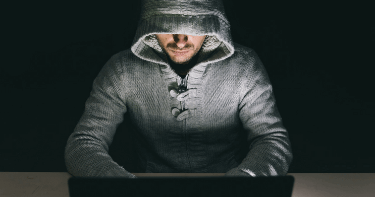 Man in a hooded sweatshirt sitting in front of a laptop computer in a dark room