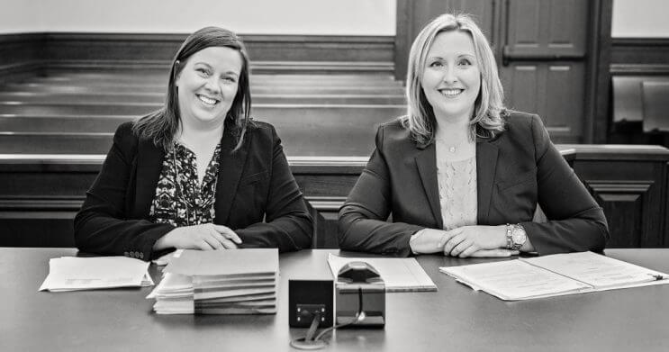 Attorney Molly Burke and Office Manager Jennifer Ford sitting at the front of an empty court room, smiling at the camera