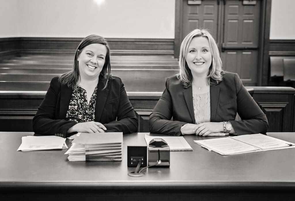 Attorney Molly Burke and Office Manager Jennifer Ford sitting at the front of an empty court room, smiling at the camera